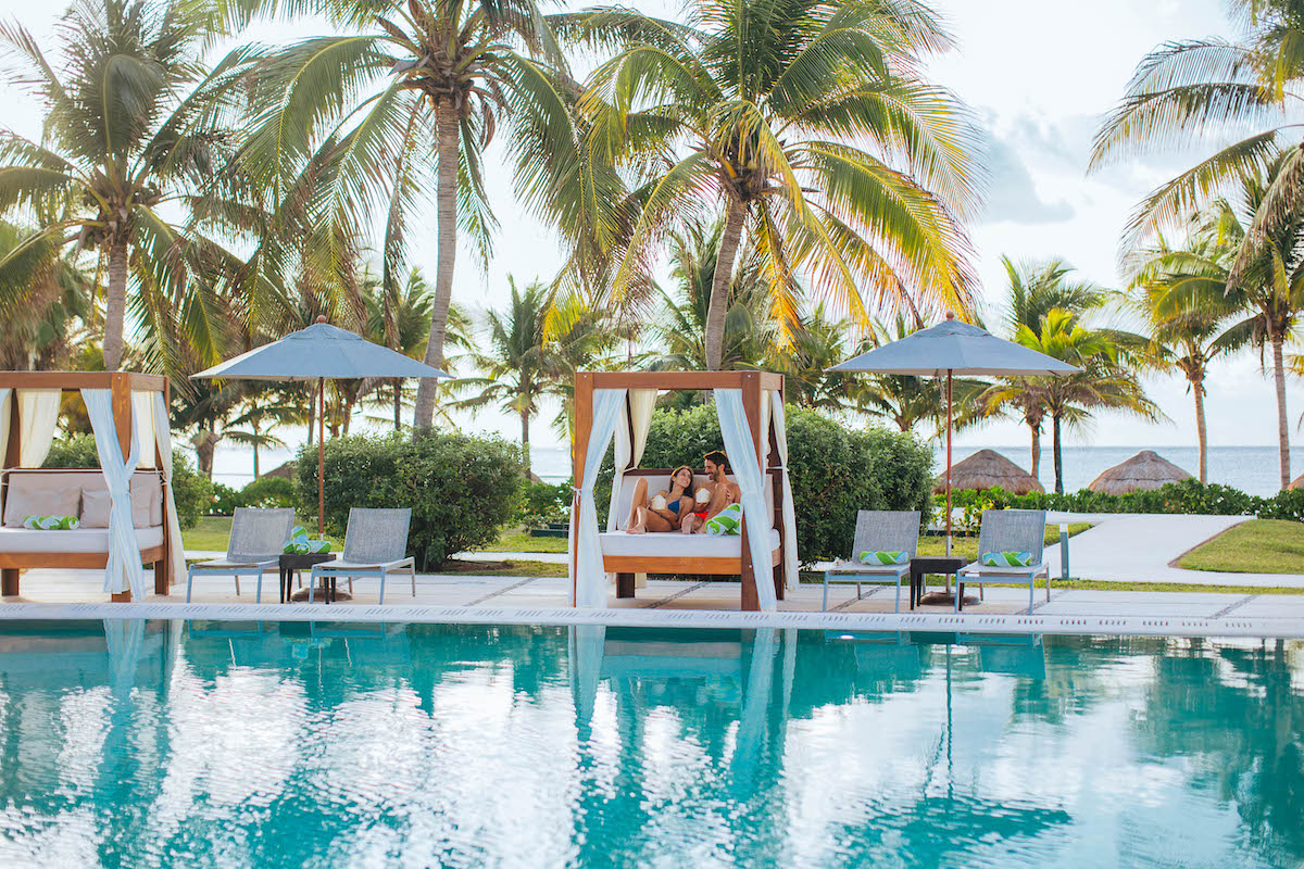 A serene outdoor pool area with cabanas, lounge chairs, and palm trees. A couple is relaxing in one of the cabanas, surrounded by greenery and a view of the ocean in the background under a partly cloudy sky.