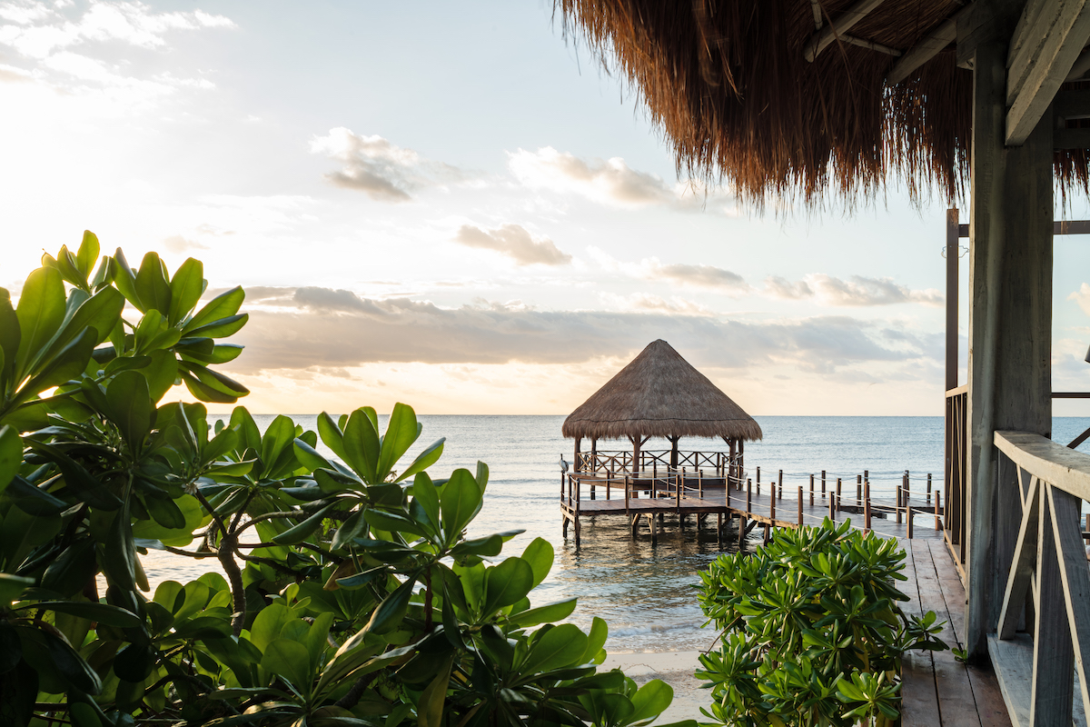 A serene coastal scene featuring a small thatched-roof hut on a wooden pier extending into calm, clear ocean waters. Lush green foliage frames the foreground, and the huts are partially seen from a viewpoint of a wooden deck at the right.