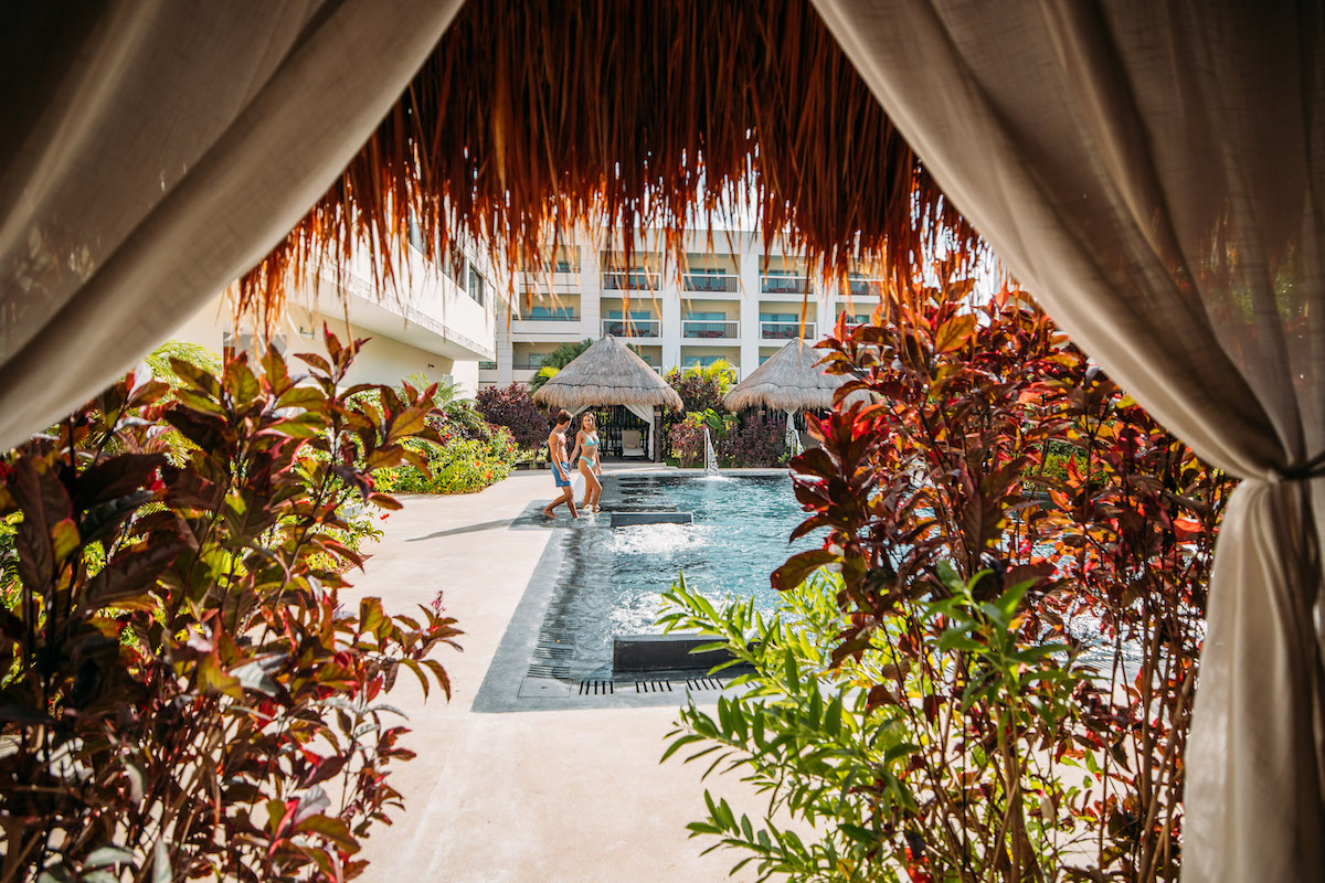 View of a resort pool framed by lush plants and white curtains, with a thatched roof structure in the background. A person is enjoying the pool near the edge with foliage on either side, providing a tropical atmosphere. Modern buildings are visible in the background.