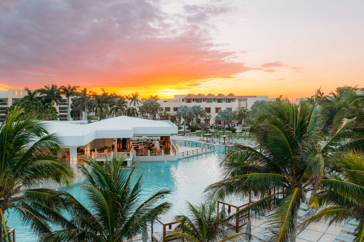 A stunning sunset over a Hyatt Ziva property in Mexico featuring a large pool surrounded by palm trees. The complex includes white modern buildings and outdoor dining areas, with the sky transitioning from vibrant oranges and purples to a soft pastel gradient.