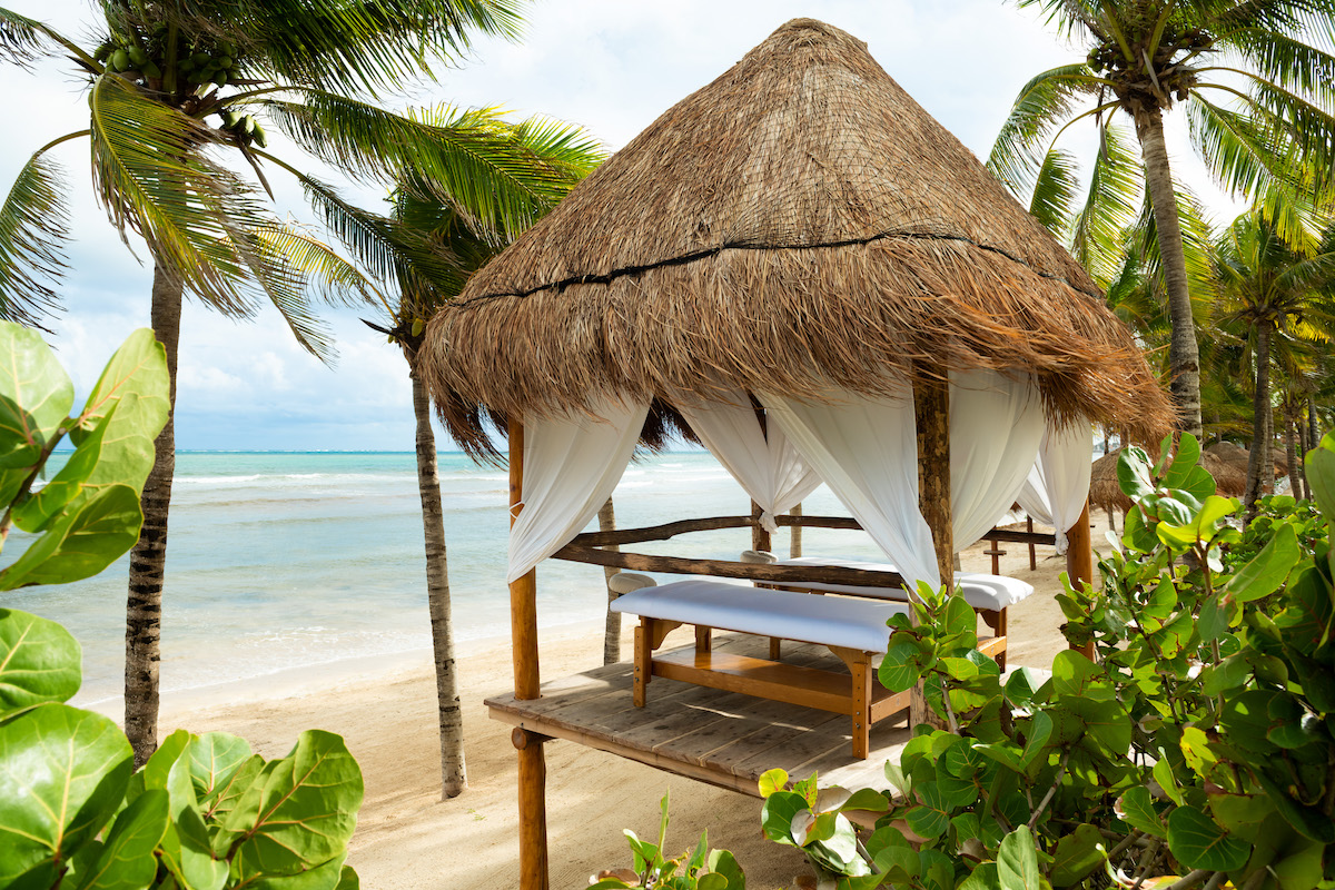 A serene beachfront scene showing a thatched-roof cabana with white curtains and a massage table inside. The cabana is set amidst palm trees and tropical greenery, overlooking a clear blue ocean under a partly cloudy sky.