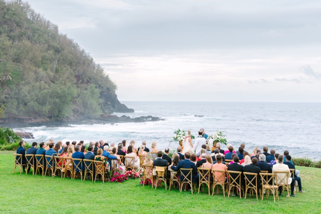 A wedding ceremony takes place outdoors near the ocean, with guests seated on wooden chairs. The background features a rocky coastline and overcast sky.