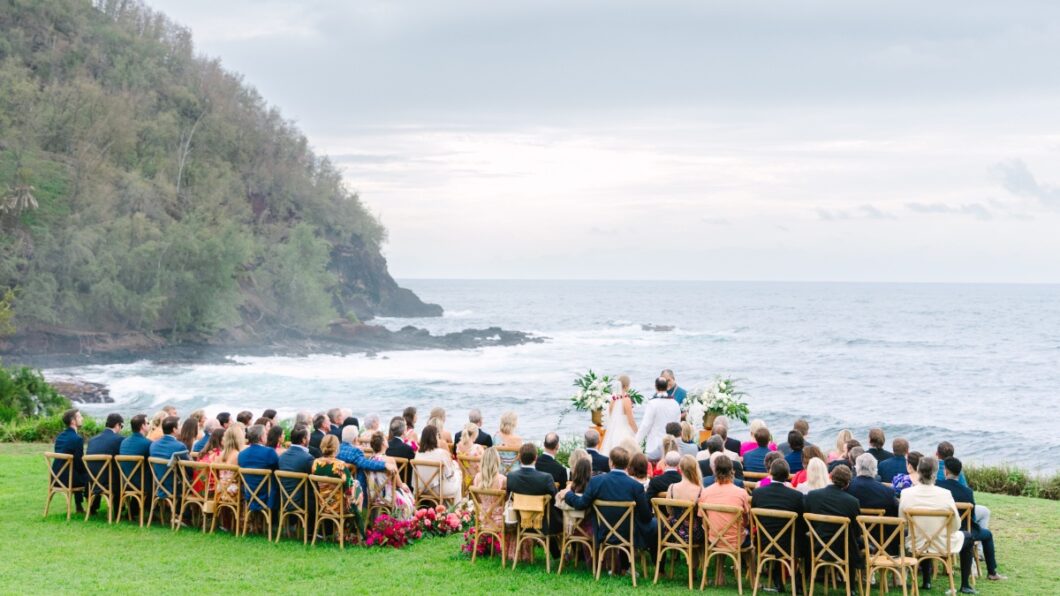 A wedding ceremony takes place outdoors near the ocean, with guests seated on wooden chairs. The background features a rocky coastline and overcast sky.