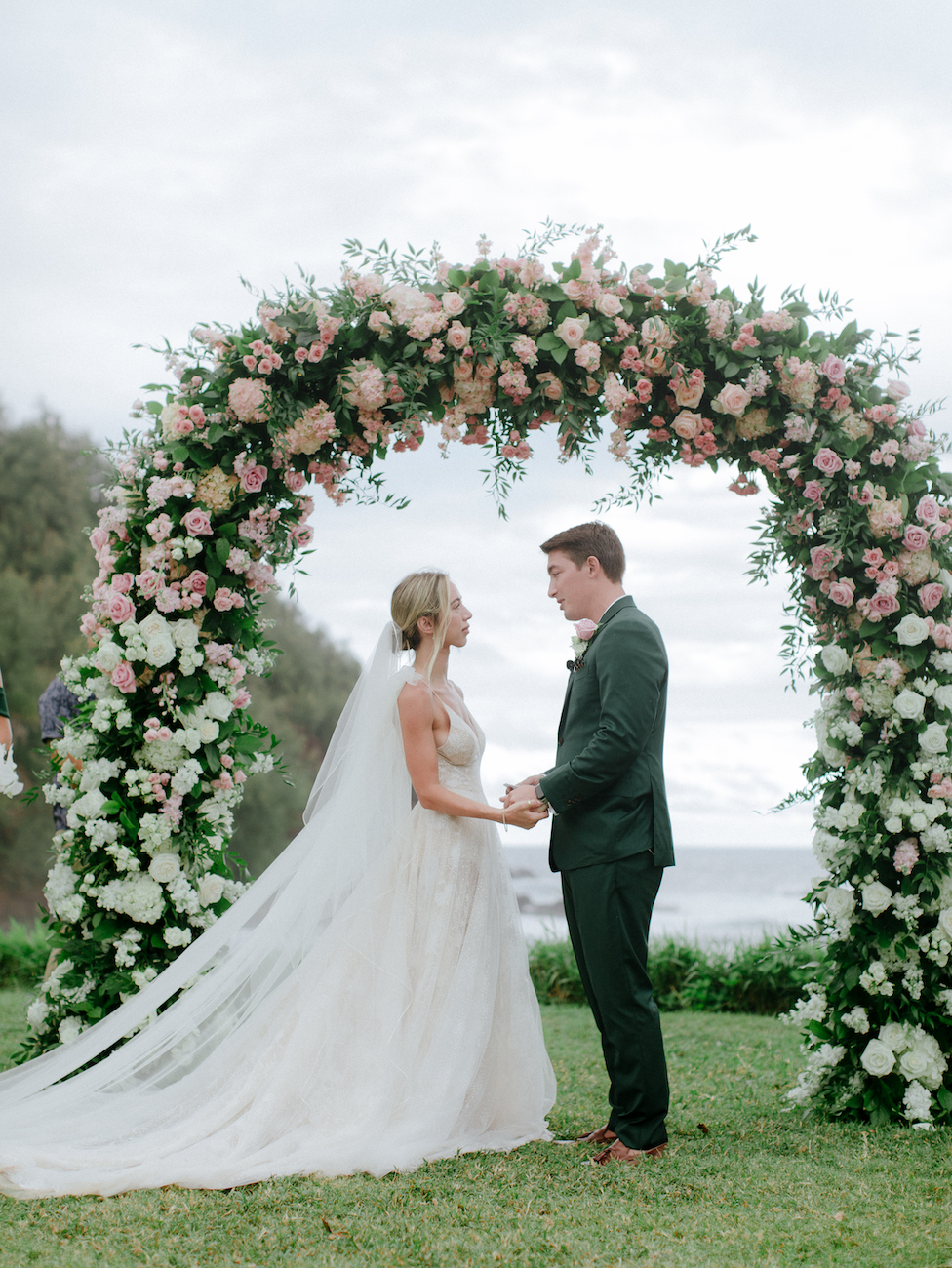 A bride and groom stand hand in hand under a floral arch during their outdoor wedding ceremony. The arch is decorated with pink and white flowers and greenery. The bride wears a long veil and a strapless gown, and the groom is dressed in a dark suit.