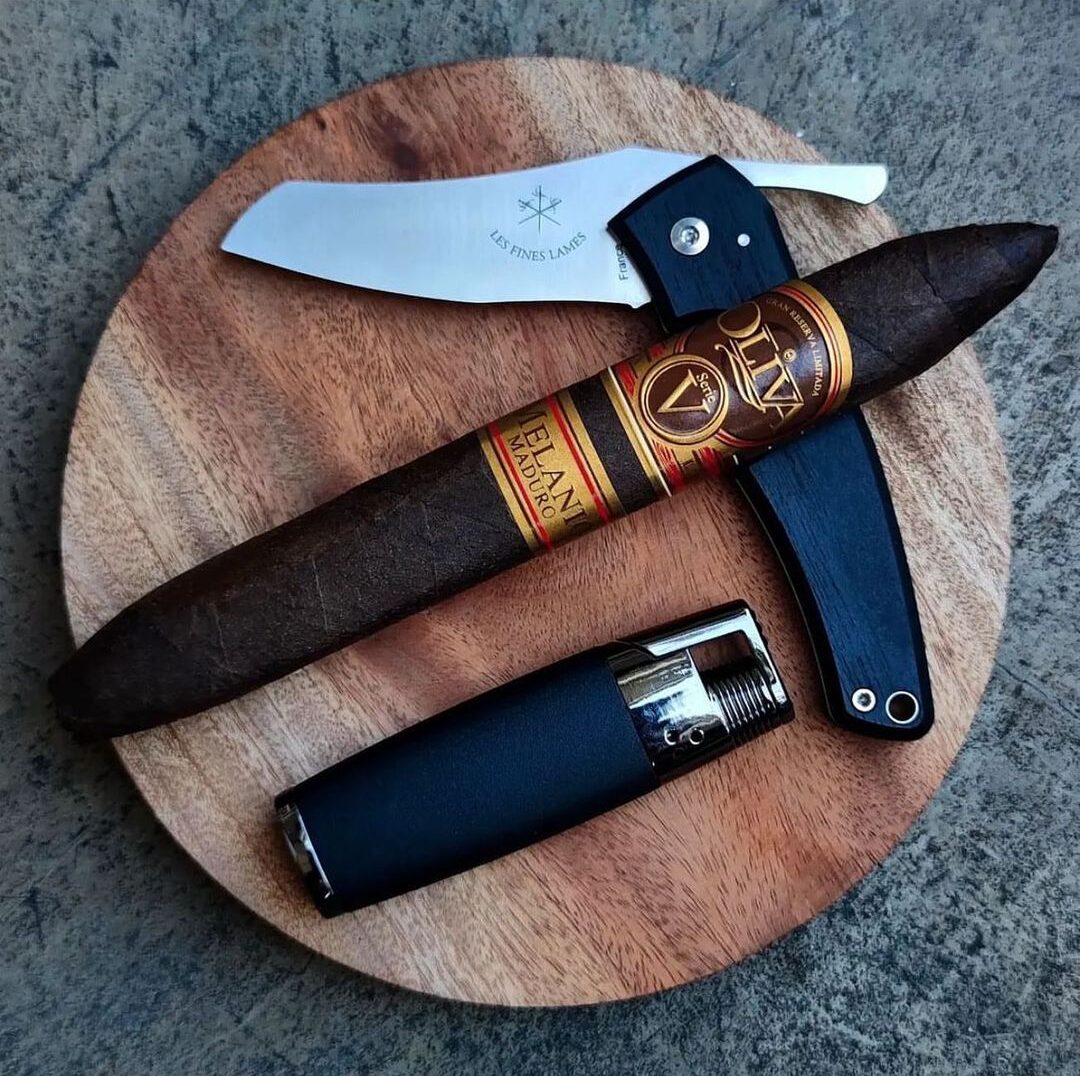 Photo of a cigar with a brown wrapper and a gold band. The cigar is resting on a wooden surface next to a silver lighter and a stainless steel knife with a black handle. 