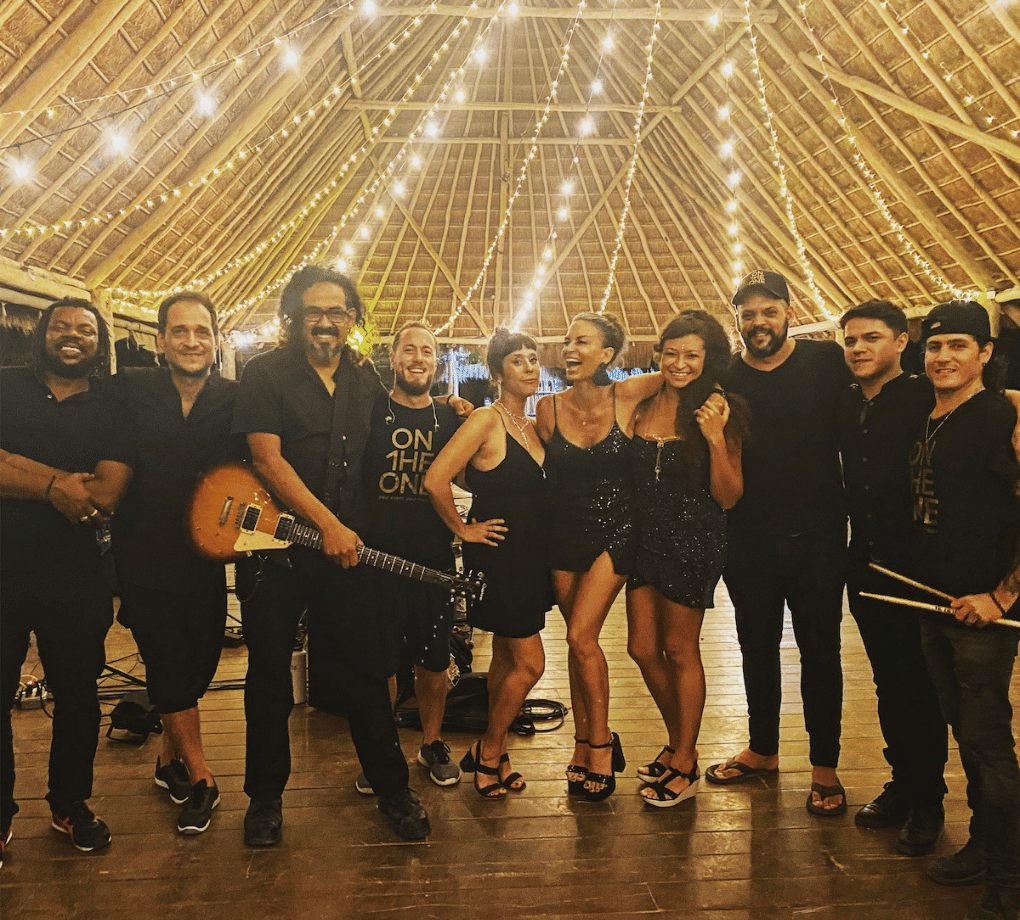 Group of people in a festive setting under string lights, with some holding musical instruments, posing on a wooden floor.