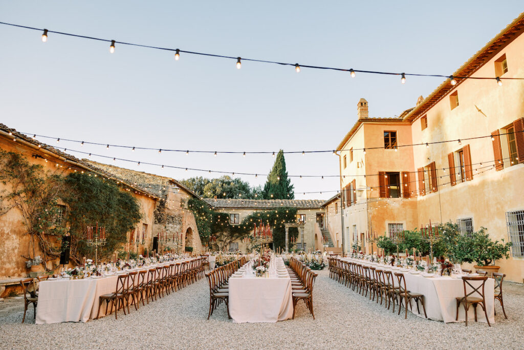 An outdoor wedding reception is set up in a charming courtyard with long tables covered in white tablecloths, elegant dining settings, and flower arrangements. String lights hang overhead, and rustic buildings with warm, earthy tones surround the space.