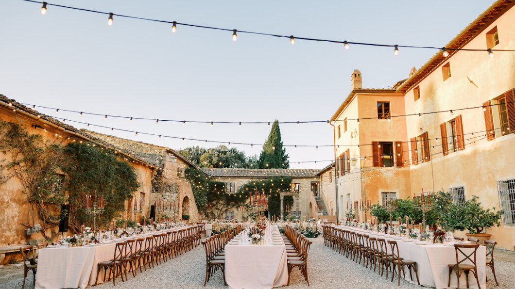 An outdoor wedding reception is set up in a charming courtyard with long tables covered in white tablecloths, elegant dining settings, and flower arrangements. String lights hang overhead, and rustic buildings with warm, earthy tones surround the space.