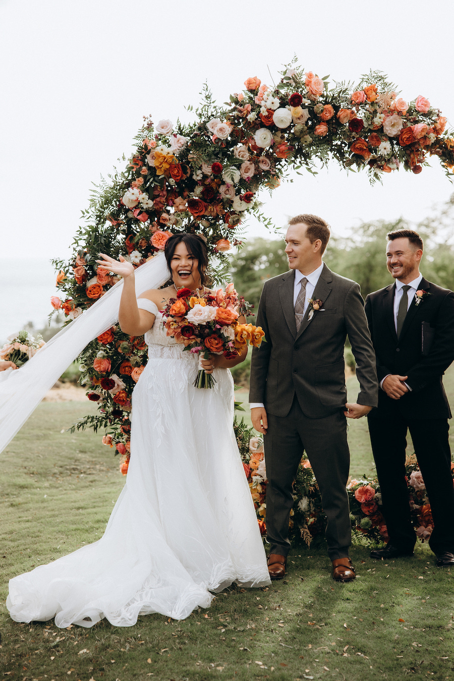 A bride in a white wedding dress, holding a bouquet of flowers, smiles joyfully under a floral arch adorned with orange and white blooms. She stands beside the groom in a gray suit and another man in a black suit, both smiling. The background features green trees.