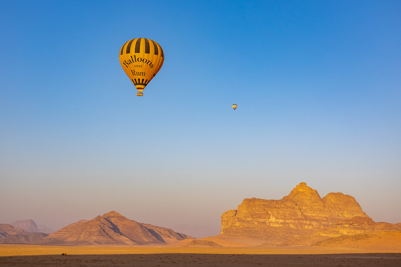 A large hot air balloon with "Balloons Over Rum" written on it flies over a vast desert landscape with sand dunes and rocky mountains in the distance under a clear blue sky. Another hot air balloon is visible in the background.