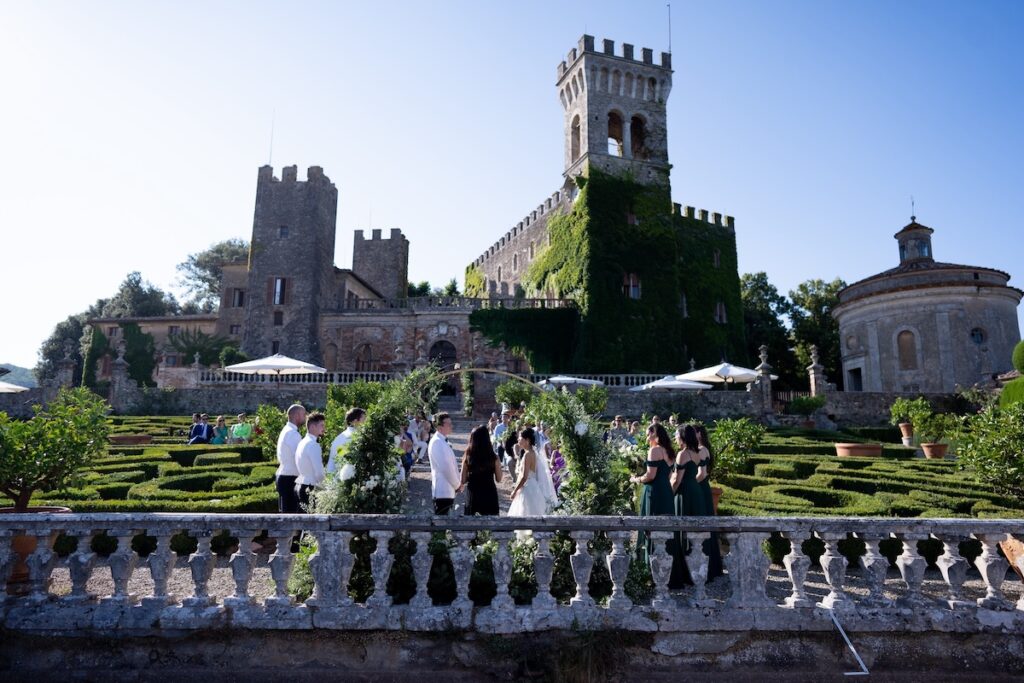 A group of people are gathered outdoors at a wedding ceremony, standing in front of a large, ivy-covered castle with turrets. The setting includes well-manicured gardens and hedges, with clear blue skies in the background. Guests are wearing formal attire.