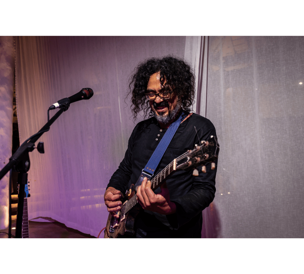 A person with curly hair and glasses is playing an electric guitar on stage. A microphone is nearby, and a curtain backdrop is visible.