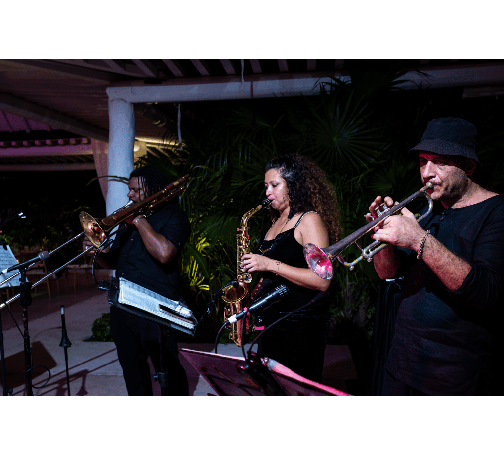 A trio of musicians playing brass and wind instruments, including a trombone, saxophone, and trumpet, perform at an outdoor venue with lush greenery in the background.