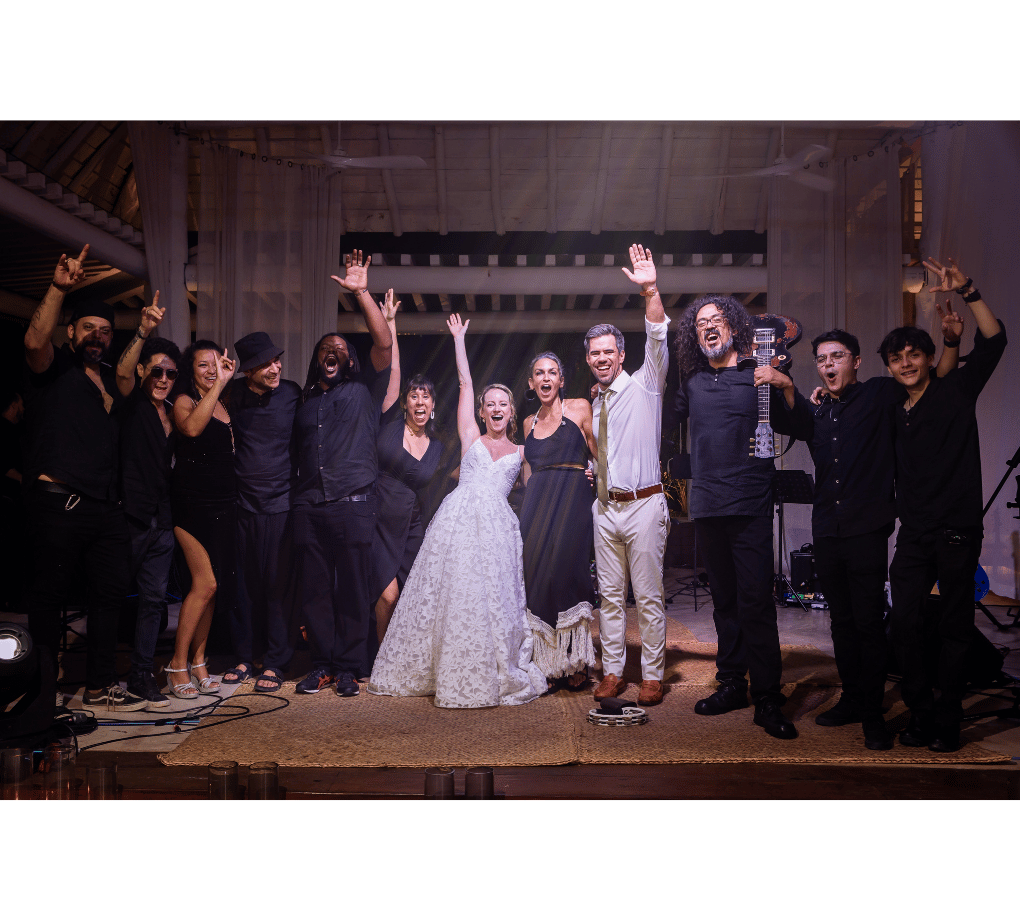 Group celebrating at a wedding. The bride in a white dress and others in black attire raise their arms, smiling on a dimly lit stage.