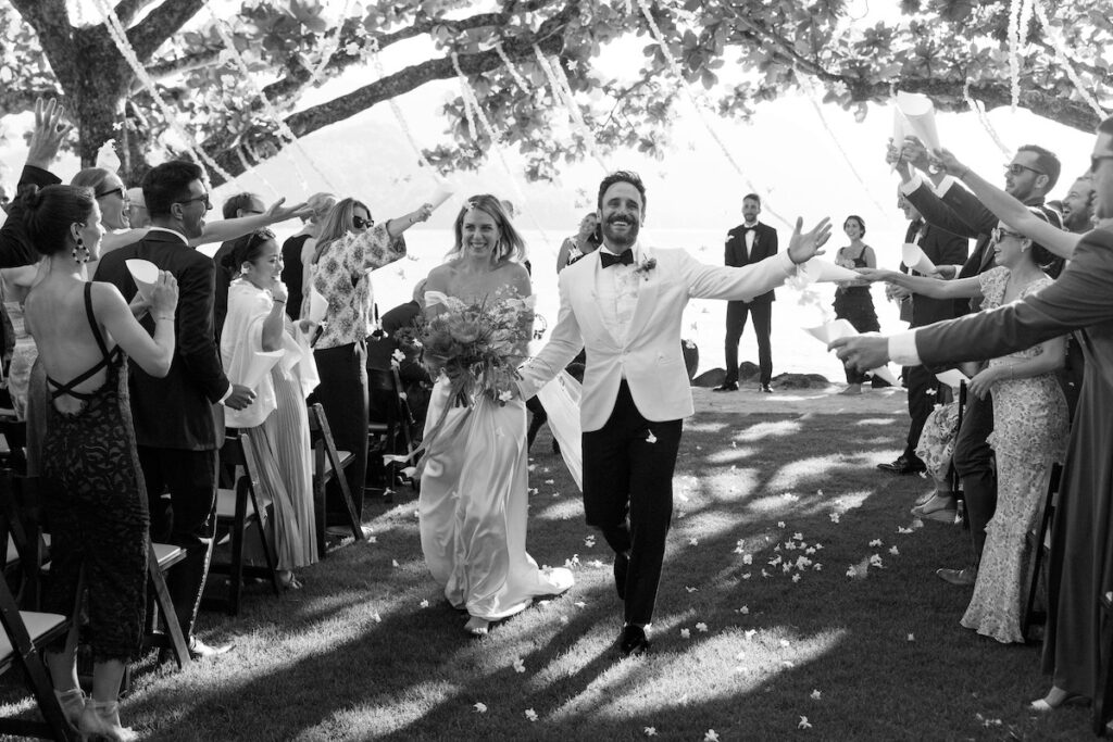 A newlywed couple walks down the aisle while guests cheer and toss petals. The event takes place outdoors under a tree canopy.