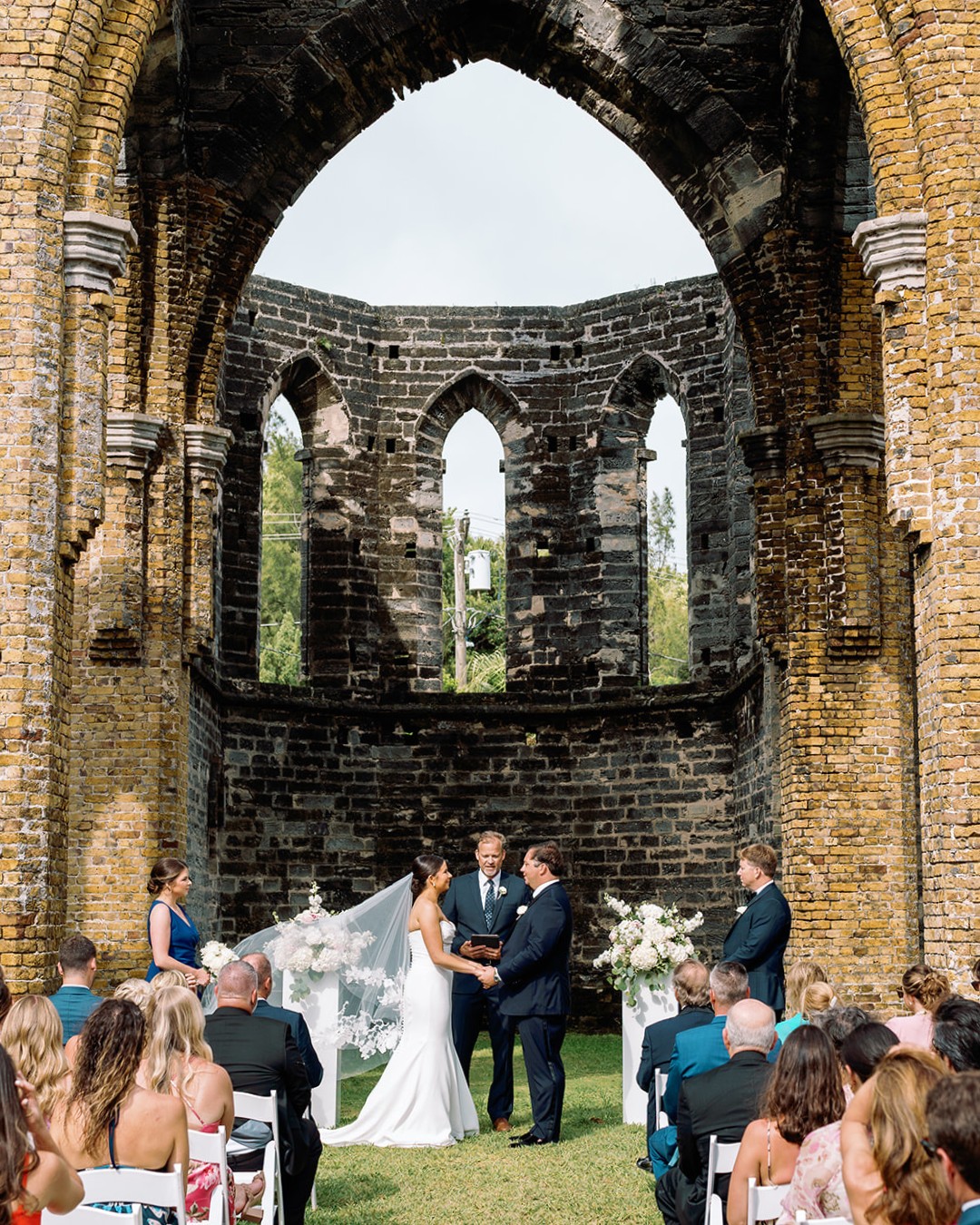 A wedding ceremony taking place outdoors in a historical, stone ruin with tall, arched openings. The bride and groom stand facing each other at the altar, surrounded by the bridal party and seated guests. The backdrop shows greenery outside the ruins.