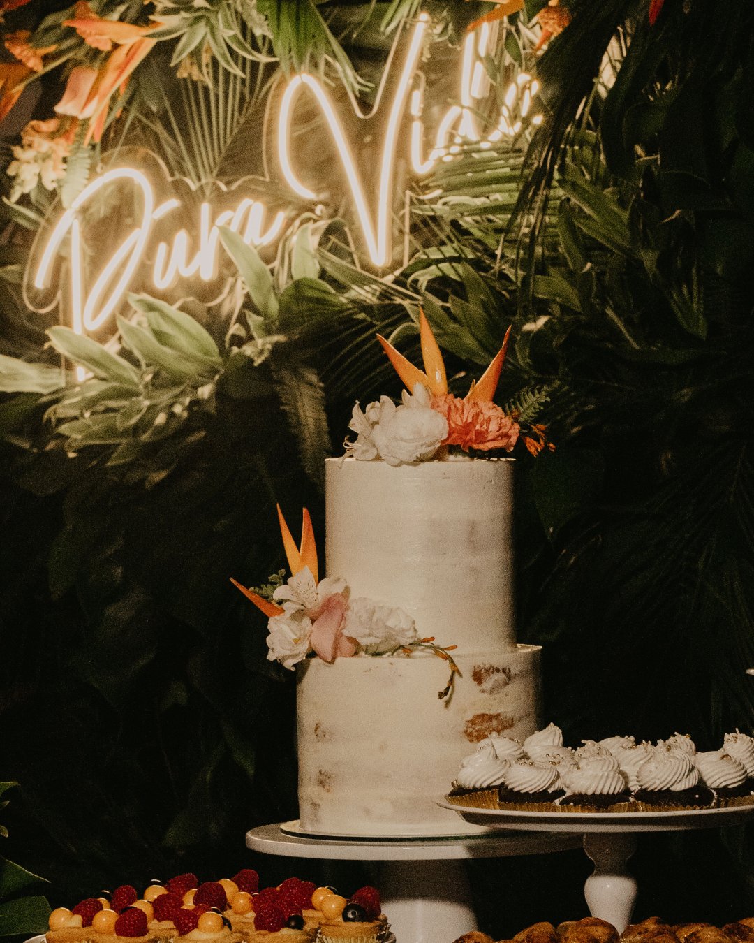 A white two-tiered cake adorned with vibrant tropical flowers stands on a table surrounded by assorted pastries and fresh fruits. Lush green foliage forms the background with a neon sign behind, partially visible with the words "Pura Vida.