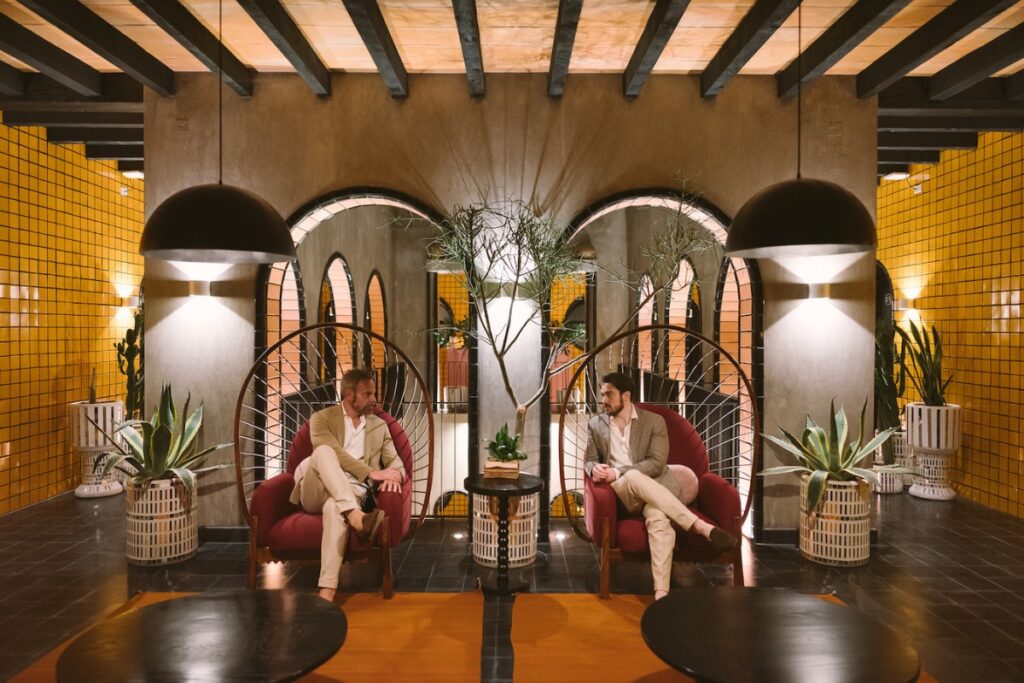 two grooms sit in boho chairs next to each other in a Spanish-style interior hotel lobby