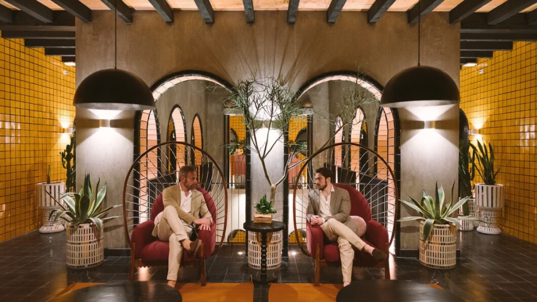 two grooms sit in boho chairs next to each other in a Spanish-style interior hotel lobby