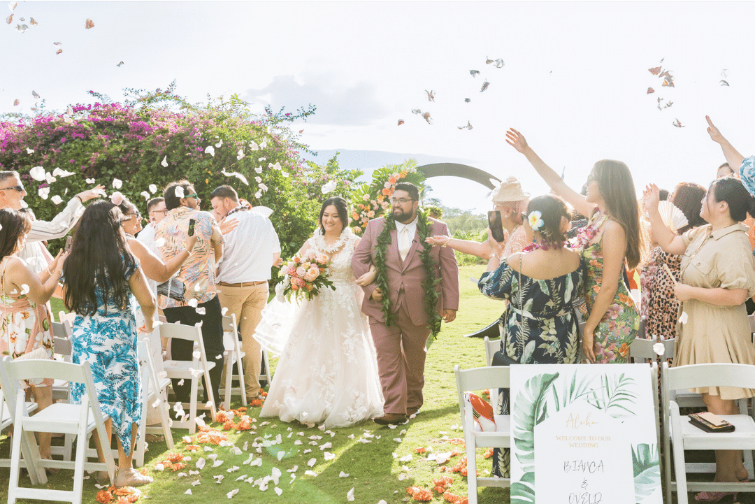 A newlywed couple walks down the aisle outdoors, surrounded by friends and family throwing flower petals. The bride wears a white gown and holds a bouquet, while the groom is in a suit. Guests, dressed in colorful attire, celebrate under a clear sky.