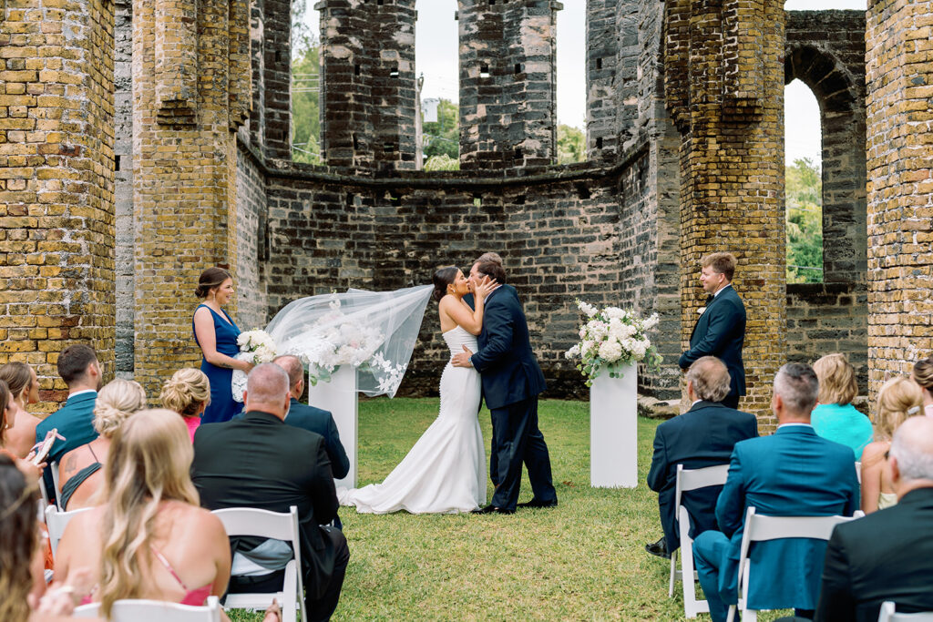 A bride and groom share a kiss at the altar under a flowing veil within the ruins of an ancient stone structure, as the bridal party and guests look on. Bridesmaids in blue dresses hold white bouquets, adding a touch of elegance to the rustic setting.