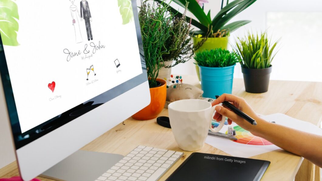 person sits at desk looking at wedding website on large desktop screen with coffee and plants sitting on the desk to the right