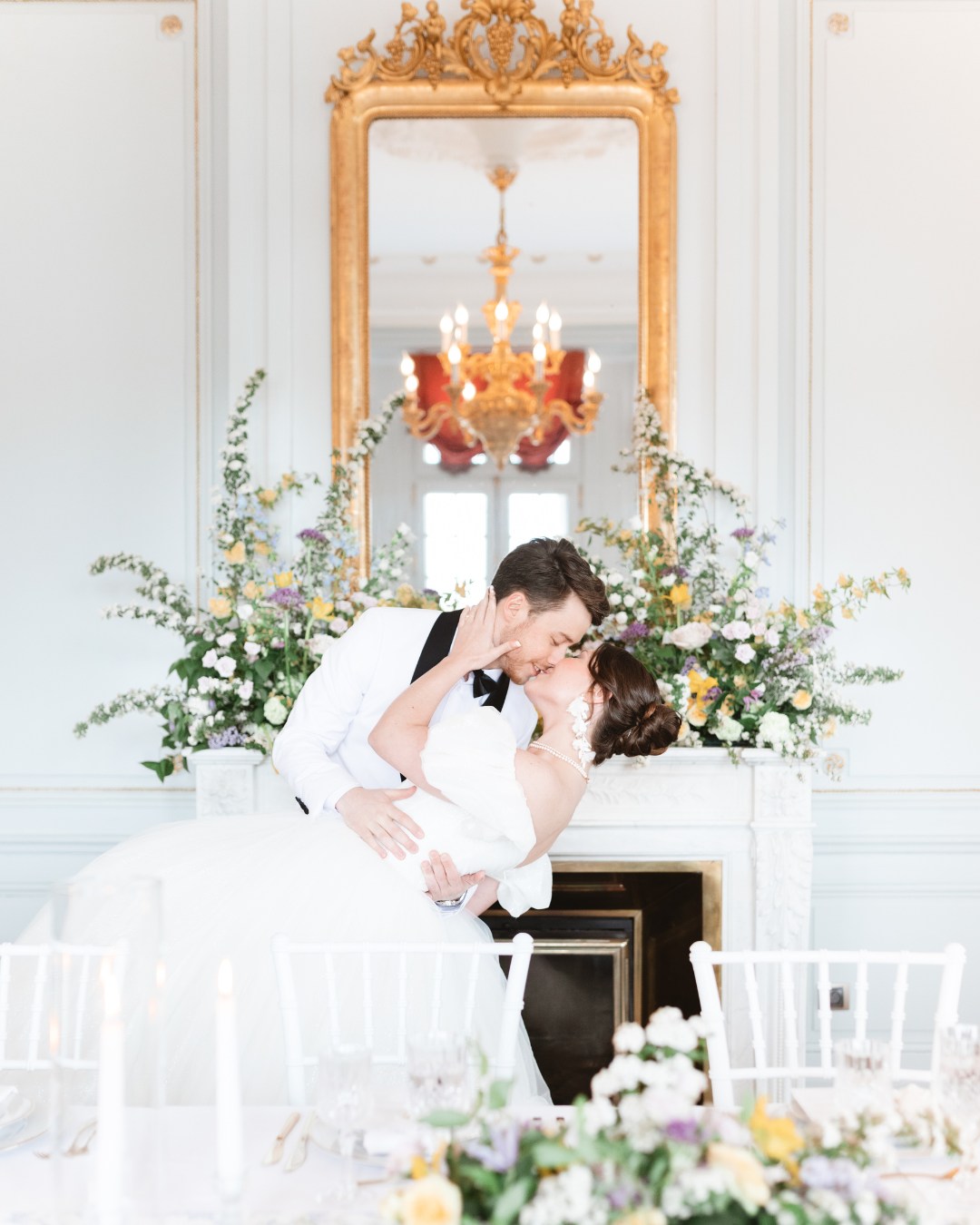 A couple dressed in wedding attire share a kiss in front of a fireplace adorned with a flower arrangement, under a large ornate mirror in an elegantly decorated room.