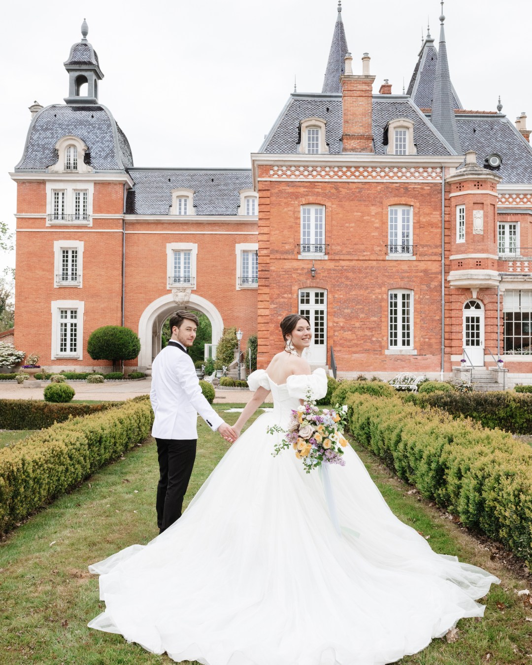 A bride and groom stand holding hands in front of a large, ornate brick building with tall, pointed roofs. The bride holds a bouquet of flowers, and the groom wears a white jacket and black pants.