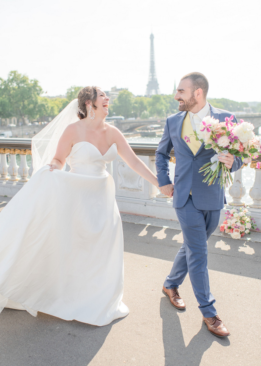 A bride in a white dress and a groom in a blue suit walk hand in hand on a bridge with the Eiffel Tower in the background. The groom holds a colorful bouquet