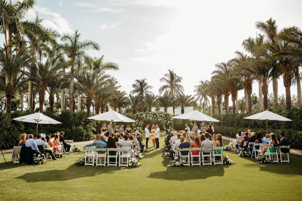 A wedding ceremony is taking place outdoors on a sunny day, with guests seated on white chairs arranged in a circular pattern around the couple. Palm trees and white umbrellas are in the background.