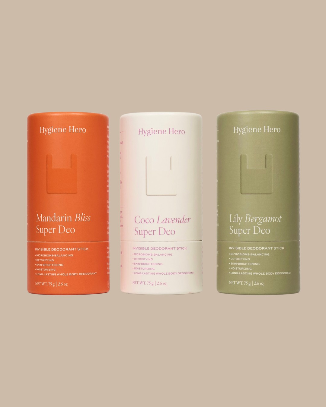 Three cylindrical deodorant sticks labeled "Hygiene Hero Super Deo" in Mandarin Bliss, Coco Lavender, and Lily Bergamot variants are standing side by side on a light brown background.