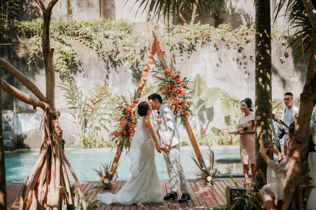 Bride and groom share a kiss at their outdoor wedding ceremony, surrounded by tropical plants and standing in front of a triangular floral arch. Guests watch and capture the moment.