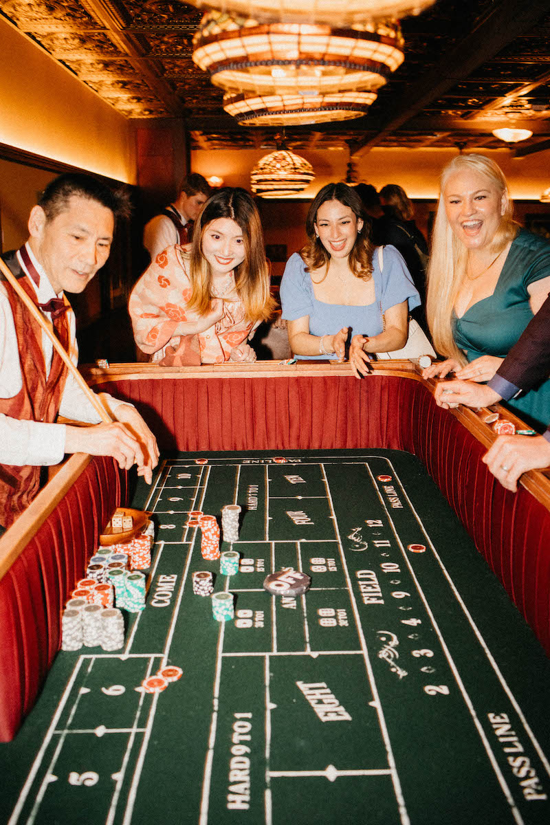 People are gathered around a craps table, engaged in a casino game, with the dealer distributing chips and dice on the table.