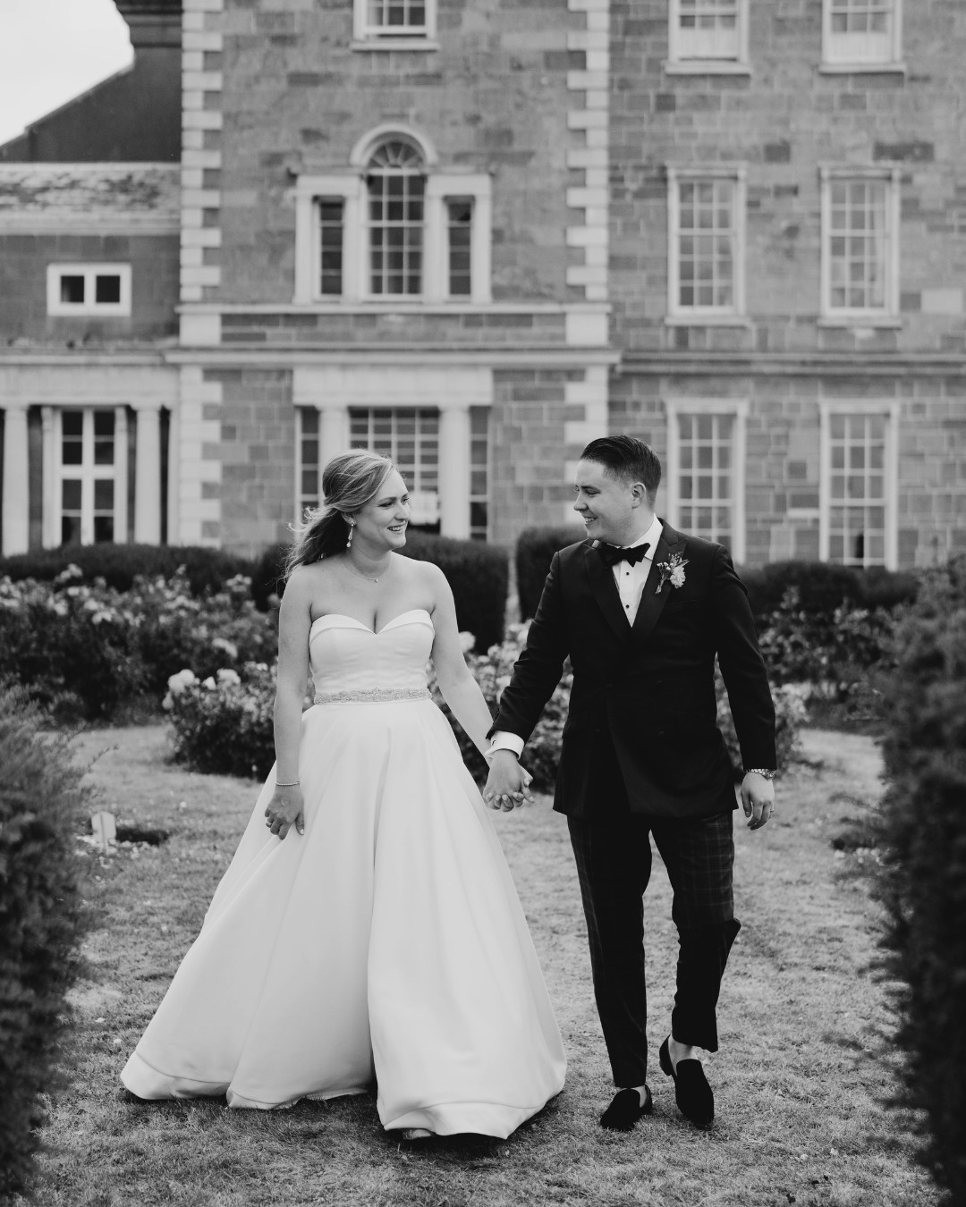 A bride and groom holding hands, both smiling, walk in front of a large historic stone building. The bride is in a strapless gown, and the groom is in a dark suit with a bow tie.