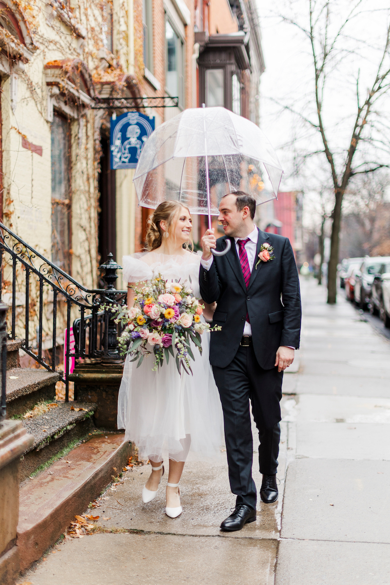 A bride and groom walk down a rainy sidewalk under a clear umbrella. The bride holds a bouquet and wears a white dress, while the groom is in a black suit with a pink tie.