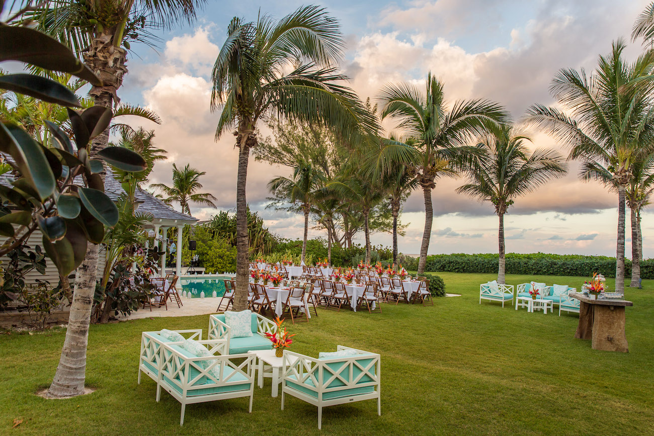 Outdoor event setup with tables and chairs arranged on a lawn surrounded by palm trees. Decorative flower arrangements are placed on the tables and nearby seating areas, with a building in the background.