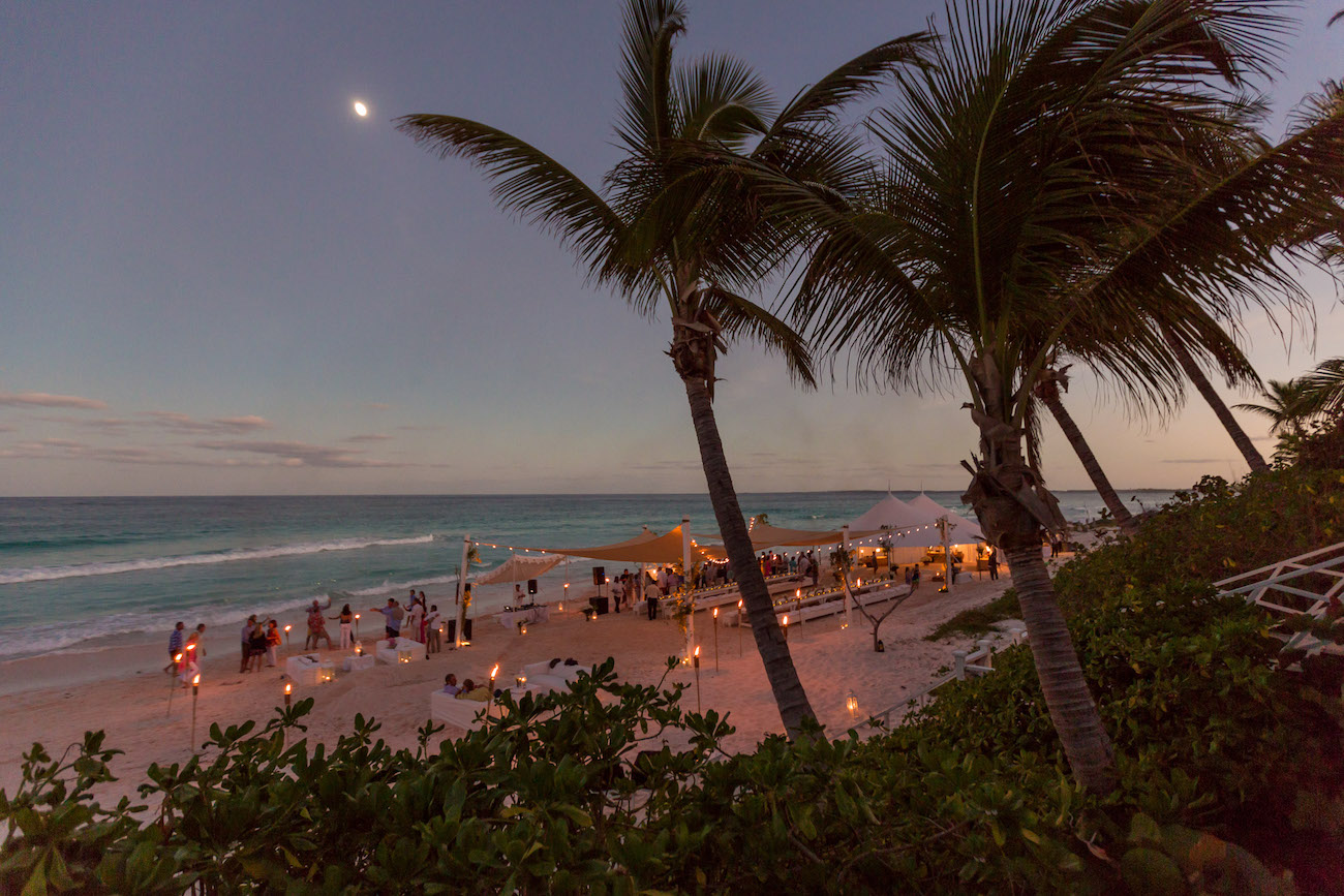 A beach scene at dusk with palm trees, tents, and numerous people gathered under string lights. The sky is clear with a crescent moon visible.