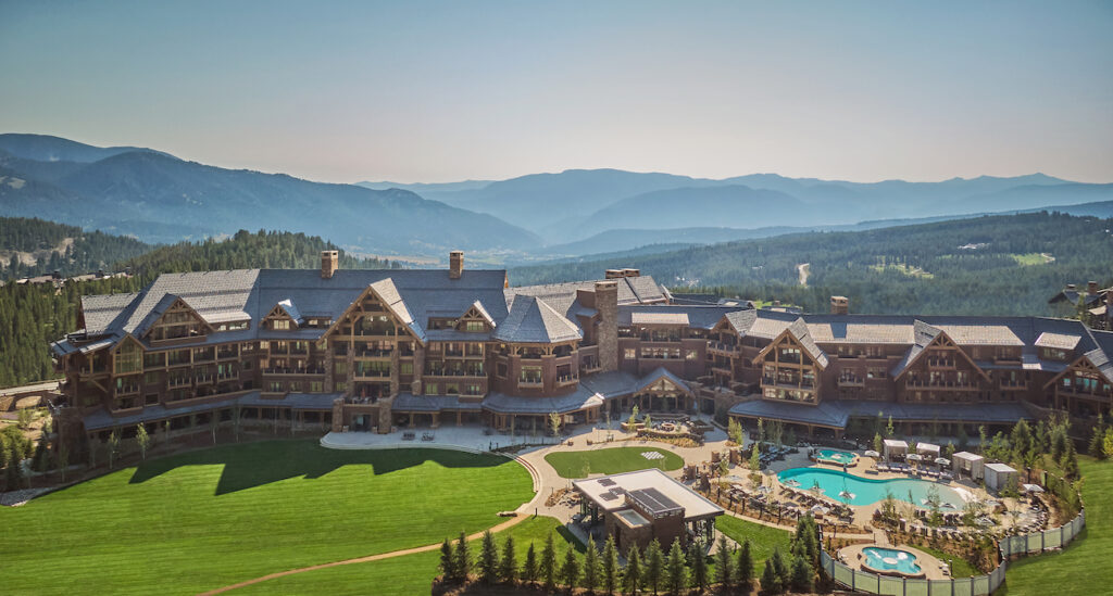 Aerial view of a luxurious mountain resort with expansive greenery, multiple buildings with gabled roofs, and a large swimming pool. The resort is set against a backdrop of rolling, forested hills and distant mountains under a clear sky.