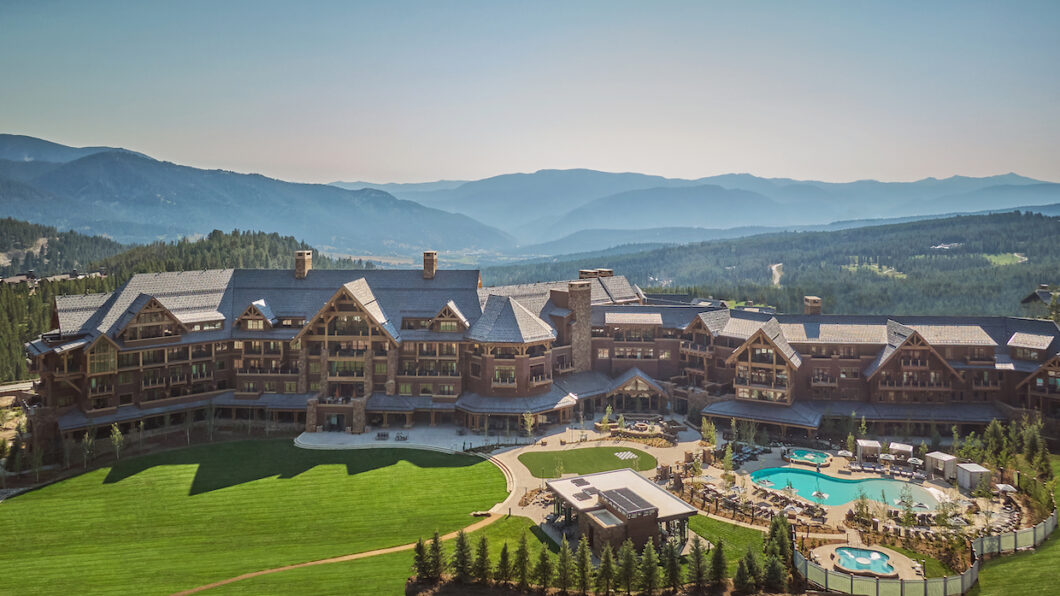 Aerial view of a luxurious mountain resort with expansive greenery, multiple buildings with gabled roofs, and a large swimming pool. The resort is set against a backdrop of rolling, forested hills and distant mountains under a clear sky.