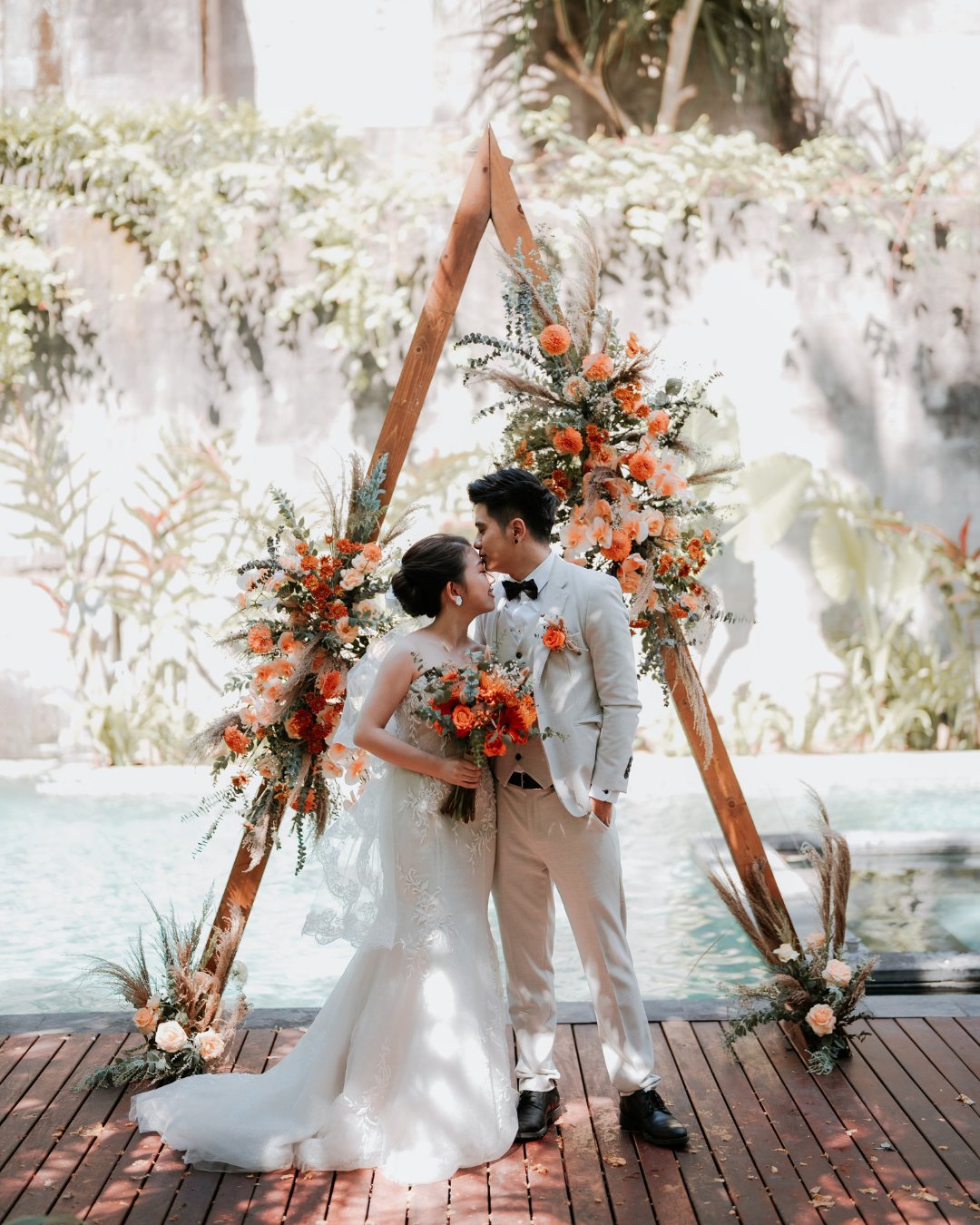 A bride and groom stand in front of a triangular floral arch by a pool, kissing. The bride holds a bouquet of orange and white flowers, and both are dressed in formal wedding attire.