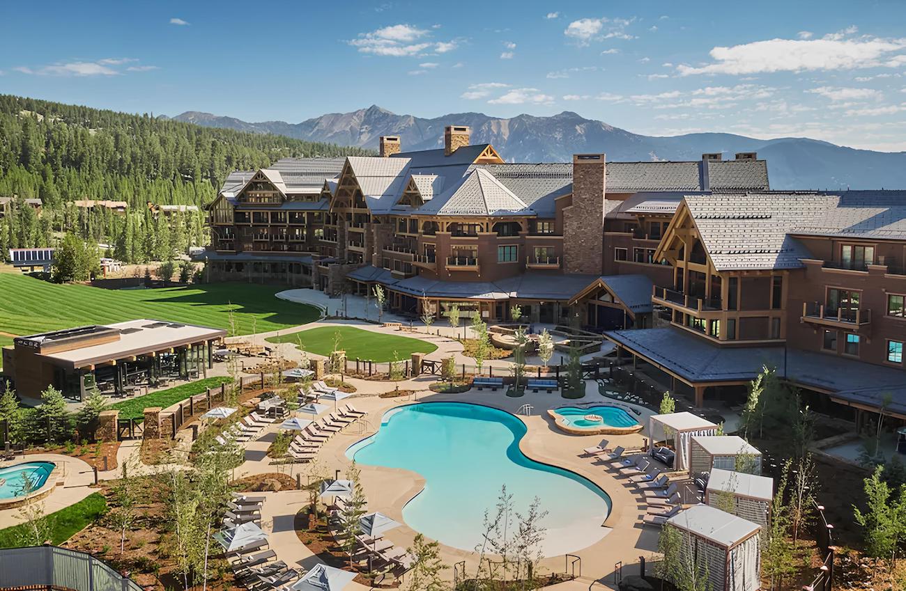 A luxurious mountain resort with an outdoor pool surrounded by lounge chairs, green lawns, and trees. The large lodge-style buildings feature wood and stone architecture. Forested mountains are in the background under a clear blue sky.