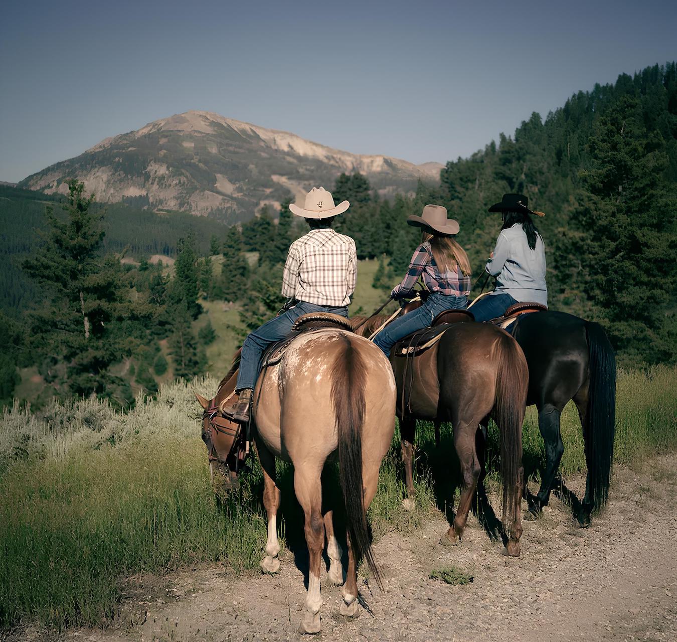 Three people dressed in plaid shirts and cowboy hats ride horses on a dirt path overlooking a scenic mountain landscape with lush greenery and trees. The sky is clear, and the group appears to be enjoying the view ahead.