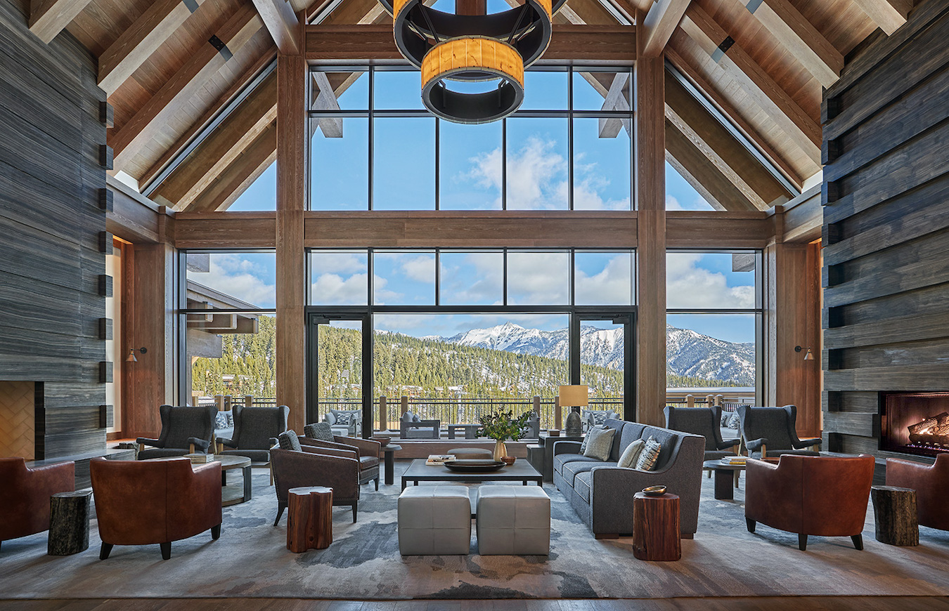 A spacious living room with large windows offering a panoramic view of snow-capped mountains. The room features wooden beams, modern furniture including sofas, armchairs, a coffee table, a chandelier, and a fireplace, creating a warm and inviting atmosphere.