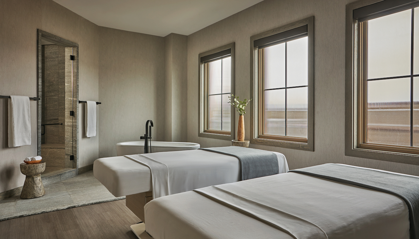 A minimalist spa room features two massage tables, each with white linens and gray blankets. A modern bathtub stands near wooden paneled windows. A small stone side table holds rolled towels beside a vase with flowers, enhancing the serene ambiance.