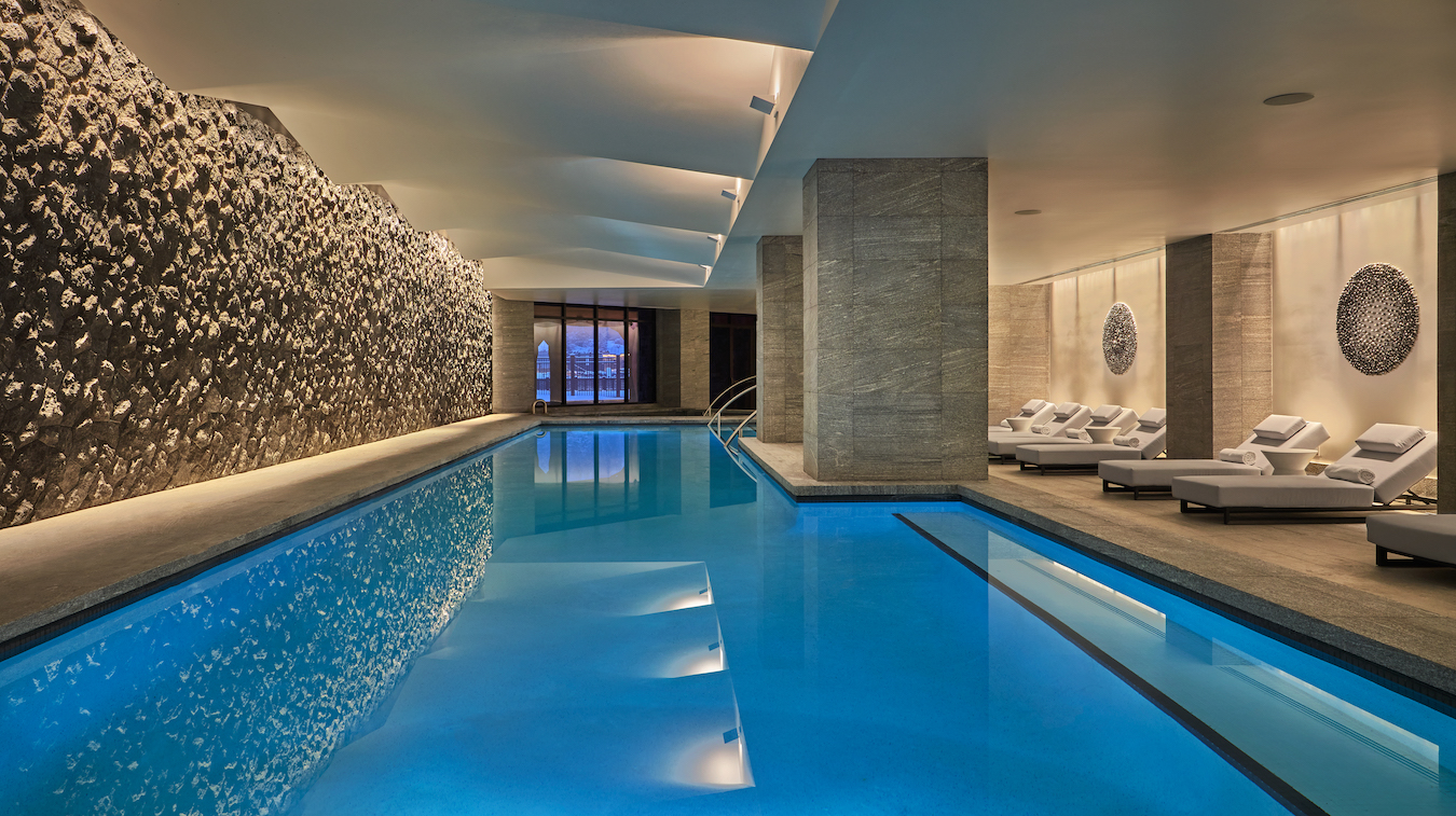 Indoor swimming pool area with a serene ambiance. The pool is illuminated with blue lighting, and the space features a wall of textured stones. On the other side, there are multiple white lounge chairs with pillows, arranged under soft, warm lighting.