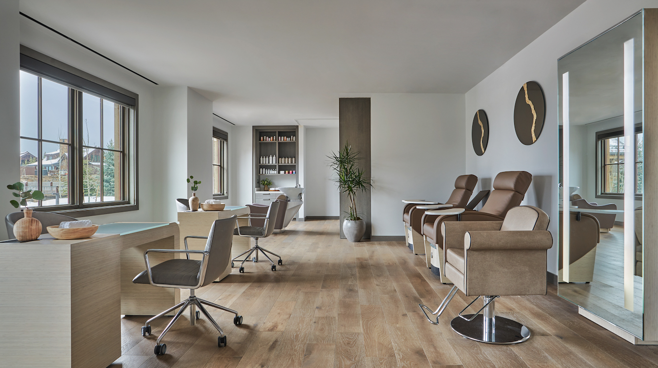 A modern, well-lit salon features wide windows, wooden floors, and minimalist decor with neutral tones. The room has three salon chairs, desk with chairs, a tall potted plant, wall art, bookshelves, and a large mirror. Some greenery is placed on the desks and shelves.