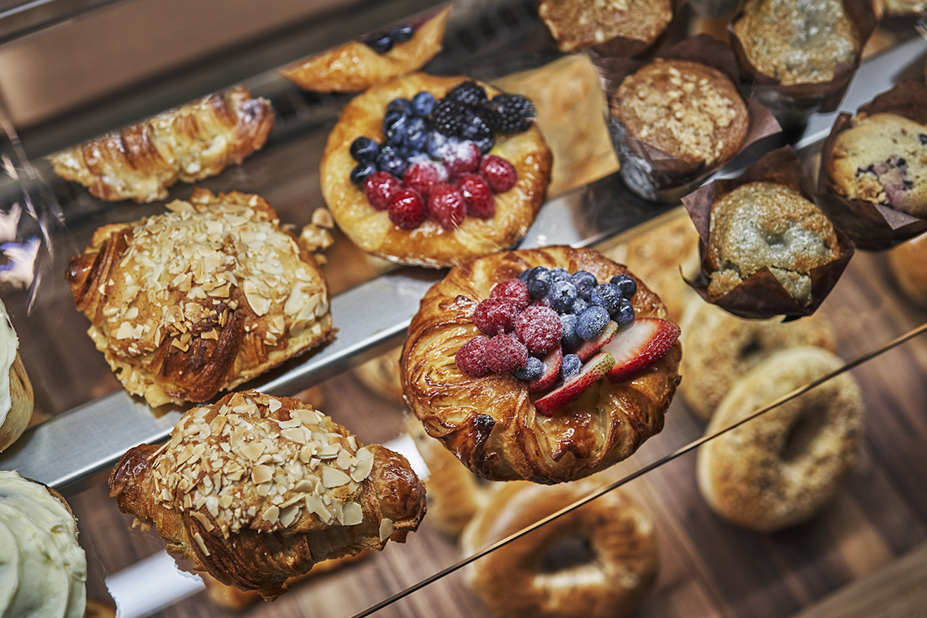 A display case filled with an assortment of pastries, including almond-topped croissants, fruit-topped danishes with strawberries, blueberries, and raspberries, muffins, bagels, and other baked goods, all arranged on wooden shelves.