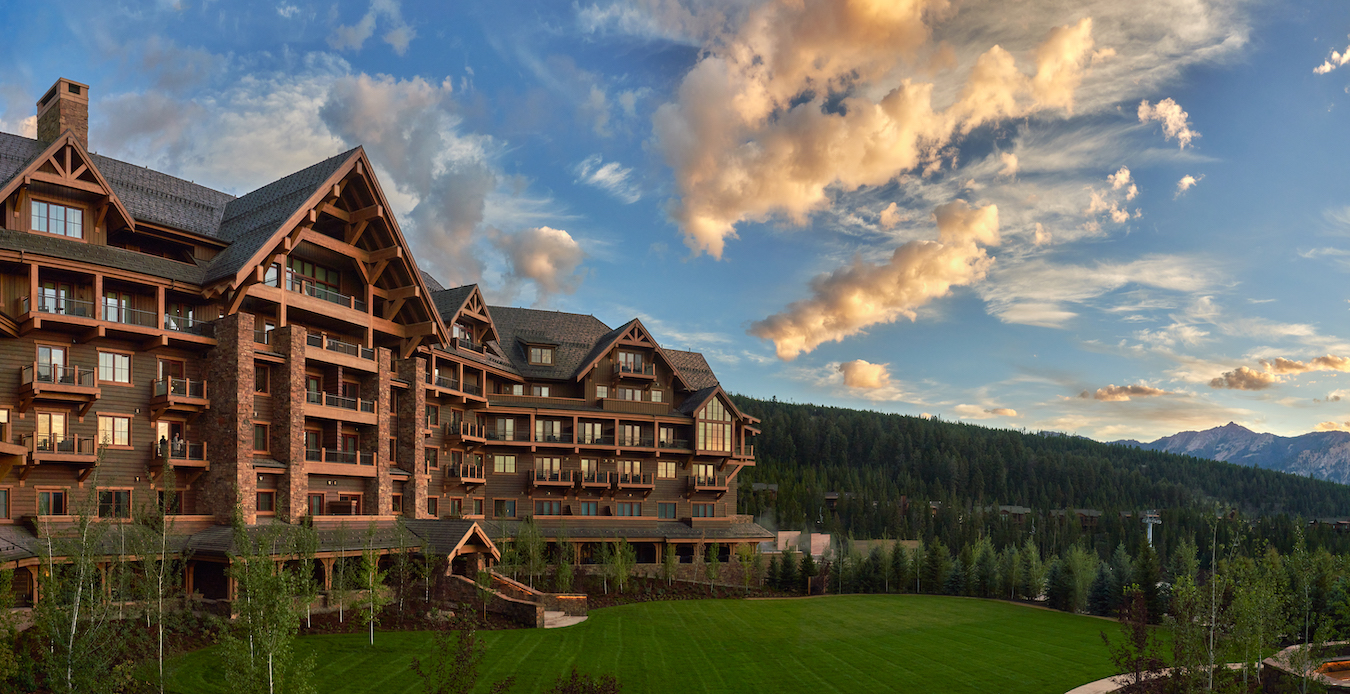 Panoramic view of a large, rustic lodge-style hotel surrounded by lush greenery and set against a backdrop of mountains and a vibrant sunset sky filled with fluffy clouds. The building features wooden balconies and large windows, enhancing its natural surroundings.