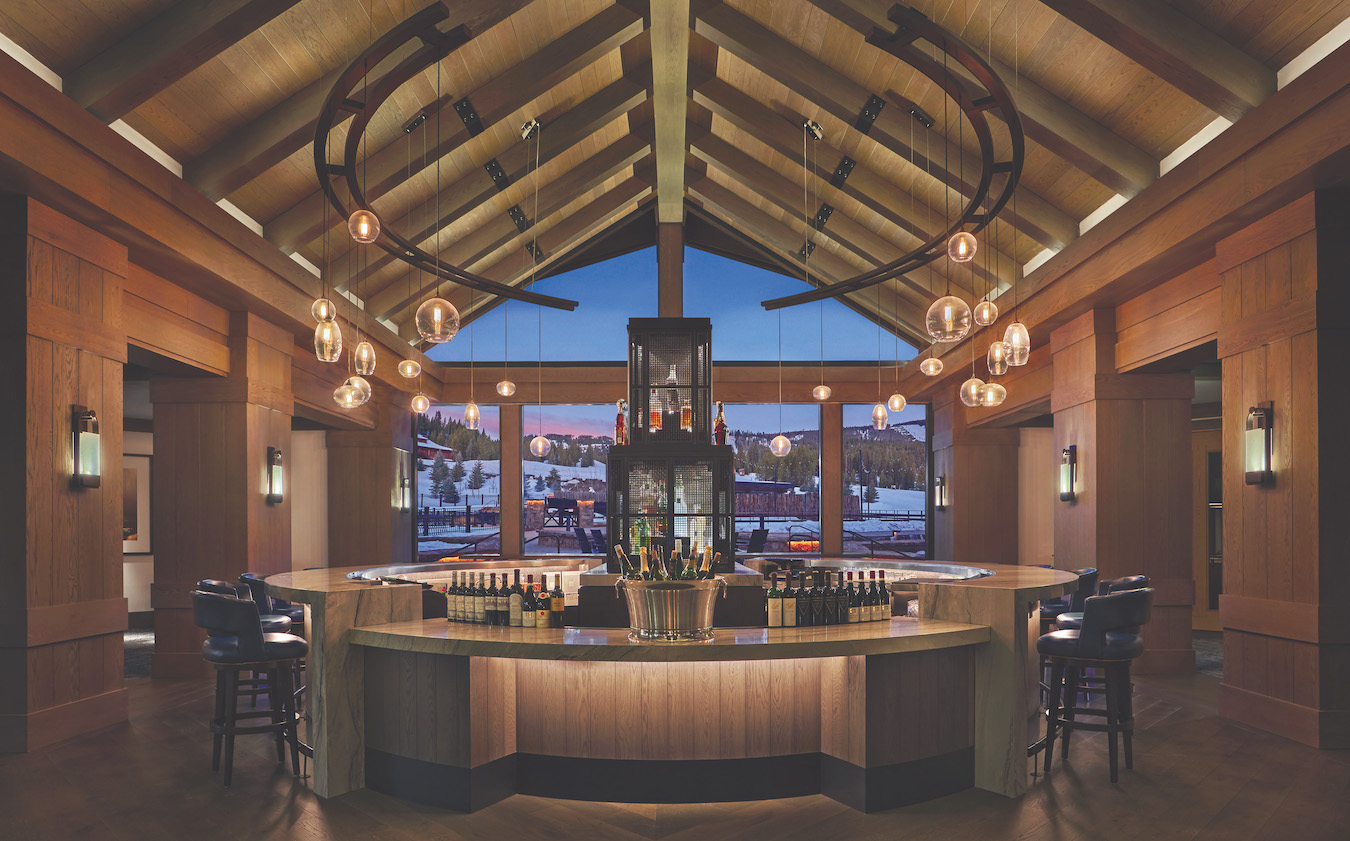 A modern bar with a wooden interior, high vaulted ceilings, and round hanging light fixtures. The curved bar counter has bottles and glasses displayed, with a view of a snowy outdoor scene visible through large windows in the background.