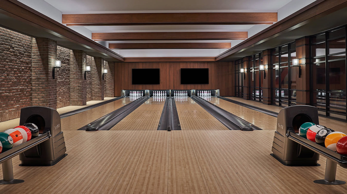 A modern bowling alley with four lanes, wooden flooring, and overhead wooden beams. Brightly colored bowling balls sit in racks on both sides near the lanes. The walls are a mix of exposed brick and wood paneling, with large windows on the right side.